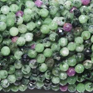 Op de afbeelding: Close-up van een streng gefacetteerde edelsteen kralen. De kralen zijn voornamelijk lichtgroen met donkerdere groene en zwarte insluitsels, en een paar magenta kralen zijn verspreid. De kralen zijn rond en hebben een gepolijst, reflecterend oppervlak.
