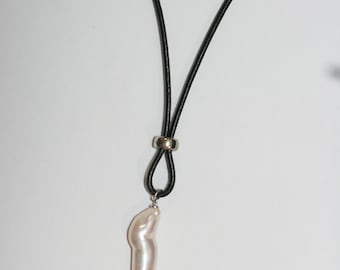Pode incluir: Um colar com um pingente de pérola barroca branca em um cordão de couro preto e um fecho prateado.