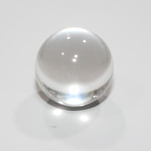 May include: A clear, transparent glass sphere. The spherical object is smooth and reflects light, creating bright spots on its surface. The sphere is sitting on a white surface.