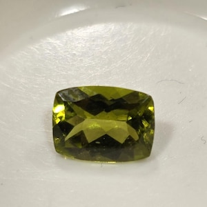 Peut inclure: Une pierre précieuse vert olive facettée, de taille coussin rectangulaire. La gemme présente une grande clarté et brillance, reflétant la lumière de multiples facettes. La pierre est posée sur un fond blanc.