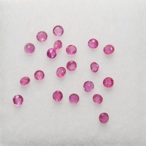 May include: A collection of pink gemstones, likely rubies, scattered on a white surface. The gemstones are round and have a smooth, polished finish.