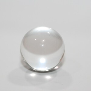 May include: A clear, spherical glass ball. The transparent orb reflects light, creating bright highlights and shadows. The smooth surface of the ball is free of imperfections, and it sits on a white surface.