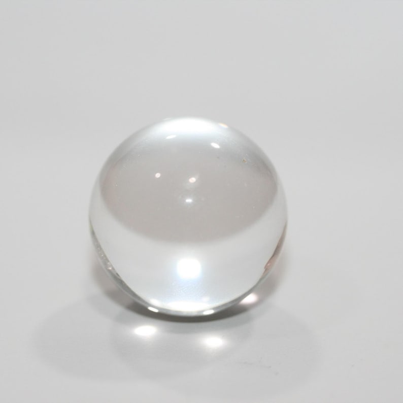 May include: A clear, spherical glass ball. The transparent orb reflects light, creating bright highlights and shadows. The smooth surface of the ball is free of imperfections, and it sits on a white surface.