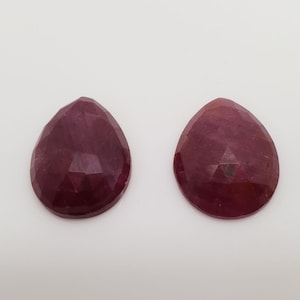Pode incluir: Duas pedras preciosas de rubi em forma de pêra com corte facetado. As pedras preciosas são de cor vermelho escuro e têm um acabamento brilhante.