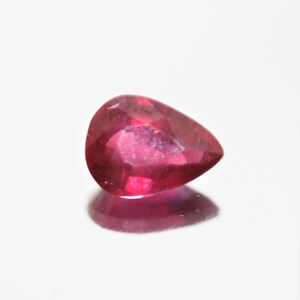 Peut inclure: Une pierre précieuse de forme poire, en rubis facetté. La gemme est d'un rouge profond et riche avec une légère lueur interne. Le rubis est posé sur un fond blanc, avec son reflet visible en dessous.