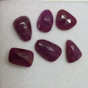 May include: Six faceted, deep red ruby gemstones of varying shapes are displayed against a white background. The gemstones include oval, rectangular, and triangular cuts, showcasing their polished surfaces and rich color.