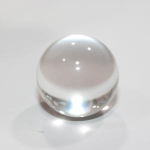 May include: A clear, spherical glass ball. The transparent orb reflects light, creating bright spots on its surface. The object is set against a white background, highlighting its smooth, rounded form. The sphere appears to be solid.
