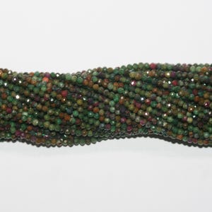 Può includere: Un filo di piccole perline sfaccettate di pietre preziose nelle tonalità del verde, del rosso e del marrone. Le perline sono rotonde e hanno una finitura lucida, creando un effetto scintillante. Le perline sono infilate insieme, formando un'esposizione strutturata e colorata.