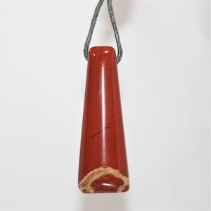 May include: A red, polished, teardrop-shaped pendant made of red jasper. The pendant has a natural, organic shape and is suspended from a gray cord.