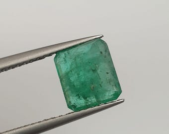 May include: A vibrant green emerald gemstone, held by silver tweezers. The emerald has a rectangular shape with faceted edges, showcasing its natural clarity and color. The gemstone is set against a plain white background.