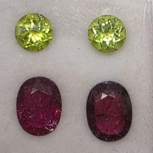 May include: Four gemstones: two round, light green peridots and two oval, deep red garnets. The gems are faceted, showing internal reflections. The peridots are a bright, clear green, while the garnets have a rich, dark red hue.