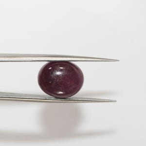May include: A small, round, polished gemstone, held by tweezers. The gem is a deep, rich burgundy color, with a smooth, reflective surface. The tweezers are silver-colored and have pointed tips. The background is plain white.