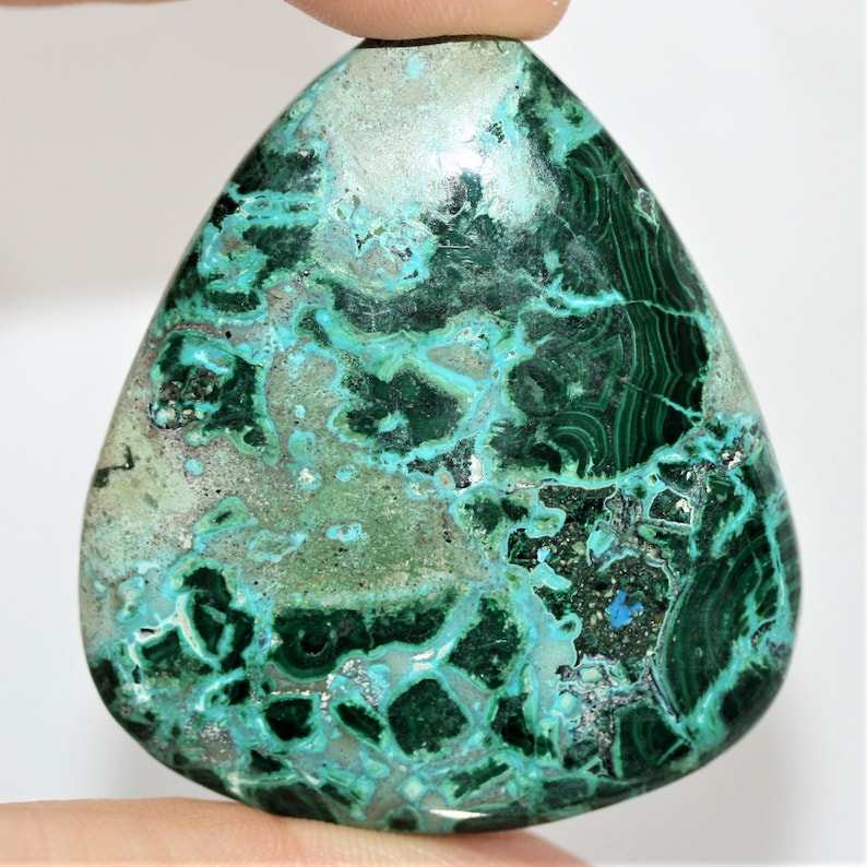 May include: A green and silver chrysocolla cabochon with a unique pattern. The stone is cut in a teardrop shape and has a smooth, polished surface.