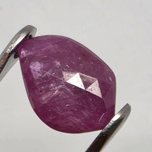 Pode incluir: Uma gema de rubi facetada, em forma oval, segurada por pinças de prata. O rubi é de uma cor rosa profunda e rica, com uma superfície polida e facetas geométricas que refletem a luz.
