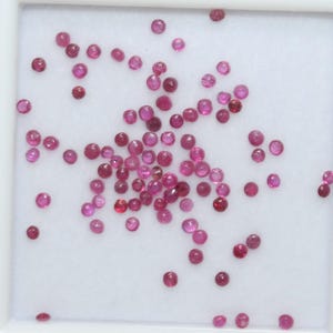 May include: A scattering of small, round, polished gemstones in shades of pink and red. The gemstones are arranged on a white surface. These gemstones could be used for jewellery making or other crafts. The gems vary in colour from light pink to deep red.