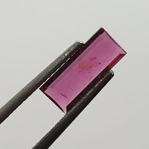 Puede incluir: Una piedra preciosa rectangular, facetada, rosa con un ligero tono púrpura. La piedra preciosa se sostiene con unas pinzas.