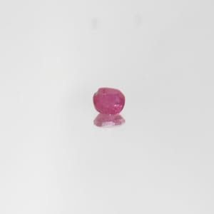 Pode incluir: Uma pequena pedra preciosa de rubi facetada em forma oval. O rubi é de cor vermelho-rosada profunda e reflete a luz, com um reflexo sutil por baixo. A pedra preciosa está sobre um fundo branco.