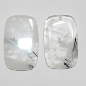May include: Two rectangular, white, translucent, cabochon-shaped stones with black inclusions. The stones have a hole drilled at the top for stringing.