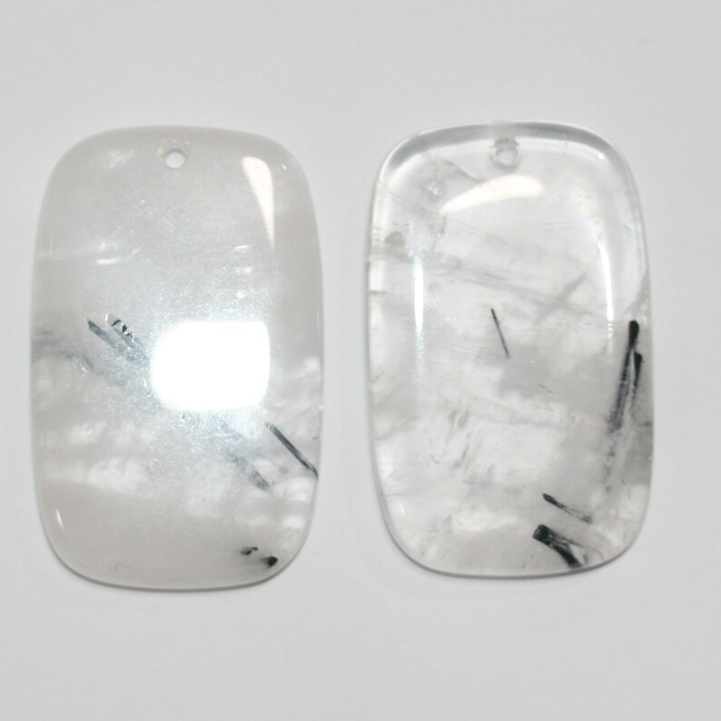 May include: Two rectangular, white, translucent, cabochon-shaped stones with black inclusions. The stones have a hole drilled at the top for stringing.