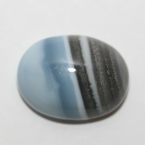 May include: A blue and gray banded opal cabochon gemstone. The opal has a smooth, polished surface and a rounded oval shape.