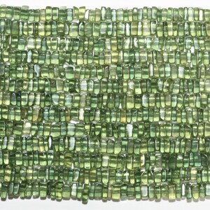 May include: Close-up of a strand of green peridot gemstone beads. The beads are rectangular with rounded edges and vary in shades of green. The beads are strung together, creating a textured surface.