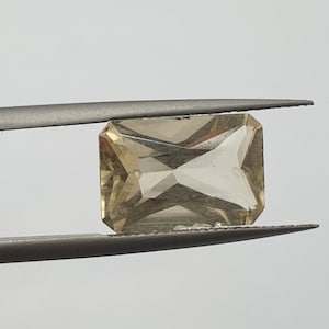 May include: A light brown, rectangular, faceted gemstone held by tweezers. The gemstone has a clear, glassy appearance.