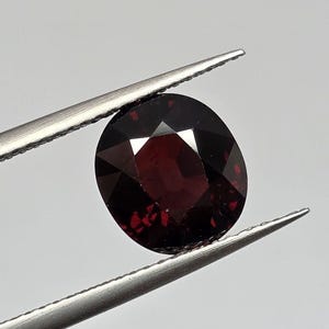 Puede incluir: Una gema facetada, de forma ovalada, de color rojo oscuro, sujeta con pinzas plateadas. La gema tiene múltiples facetas que reflejan la luz, creando un efecto brillante. La gema es de un tono rojo intenso y rico.