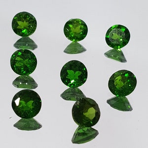 May include: Eight round, green gemstones with a star-shaped inclusion in the center. The gemstones are arranged in a scattered pattern on a white surface.