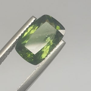 Op de afbeelding: Een groene edelsteen met een rechthoekige snede, vastgehouden door een pincet. De edelsteen is transparant en heeft een lichte glans.