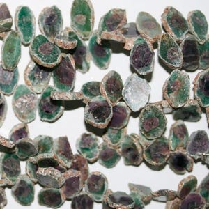 Puede incluir: Una hebra de cuentas de piedras preciosas verdes y moradas, cada cuenta tiene una forma plana e irregular con una textura rugosa y natural.