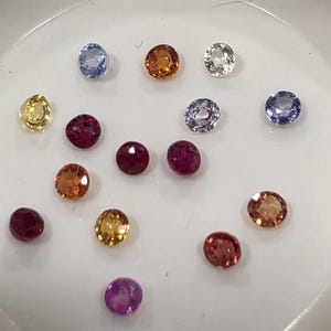 May include: A collection of loose gemstones in various colors, including yellow, orange, red, purple, and blue. The gemstones are round-cut and appear to be faceted, reflecting light. The gems are scattered on a white surface.