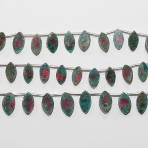 Peut inclure: Gros plan sur des perles de pierres précieuses. Chaque perle est de forme ovale allongée, avec un extérieur vert-bleu et une tache centrale rouge foncé. Les perles sont enfilées sur un fil fin de couleur argentée. Fond blanc.