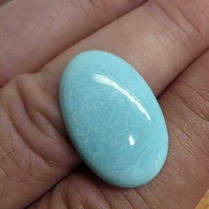 Puede incluir: Una piedra preciosa en forma de cabujón ovalado con una superficie lisa y pulida. La piedra presenta un color azul turquesa claro vibrante con sutiles inclusiones arremolinadas de color blanco y crema pálido en todo su cuerpo opaco.