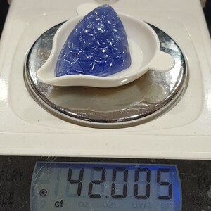May include: A blue gemstone with a faceted design is displayed on a white scale. The scale reads 42.005 ct.
