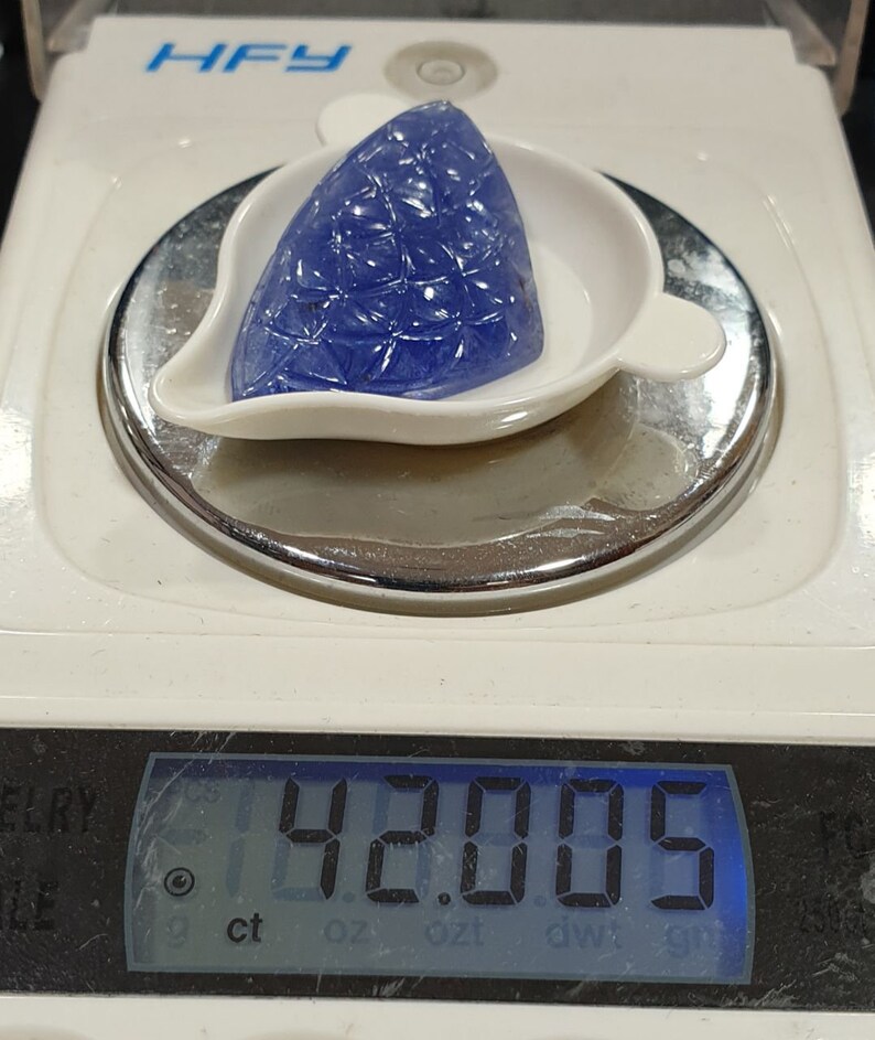May include: A blue gemstone with a faceted design is displayed on a white scale. The scale reads 42.005 ct.
