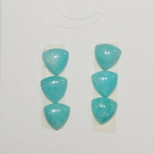 Puede incluir: Tres cabujones de color turquesa en forma de triángulo en una tarjeta blanca. Los cabujones están dispuestos verticalmente en una fila.