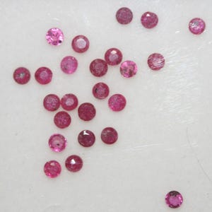May include: A scattering of small, round, faceted gemstones in shades of pink and red. The gemstones are displayed on a white surface, highlighting their polished facets and vibrant colours. These are likely rubies.