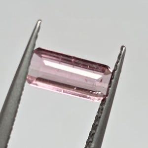 Può includere: Una gemma rettangolare rosa chiaro è tenuta da una pinzetta d'argento. La gemma appare trasparente, con linee interne e sfaccettature visibili. La pinzetta è a fuoco, evidenziando la chiarezza della gemma.
