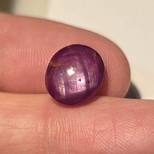 Peut inclure: Une pierre précieuse ovale polie, de couleur violet profond. La gemme présente une surface lisse avec des stries internes et un éclat subtil. La pierre est tenue entre deux doigts, mettant en valeur sa taille et sa couleur.