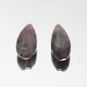 Op de afbeelding: Twee druppelvormige edelstenen met facetgeslepen oppervlakken. De stenen zijn voornamelijk grijs met vleugjes roze en rood. De edelstenen worden getoond op een reflecterend, wit oppervlak.
