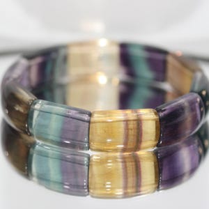 Può includere: Primo piano di un braccialetto di fluorite. Il braccialetto presenta perline rettangolari nelle tonalità del viola, verde e giallo. Le pietre lucide hanno una finitura lucida e sono infilate insieme per formare una fascia flessibile. Il braccialetto è esposto su una superficie riflettente.