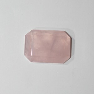 May include: A rectangular rose quartz gemstone with an emerald cut. The stone is a soft pink color and has a polished surface. The gemstone is a natural crystal, often used in jewelry and healing practices.