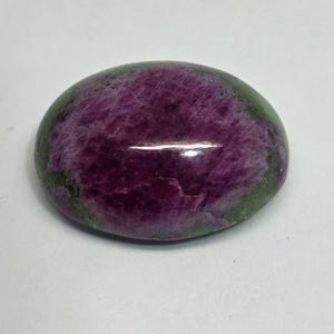 May include: Oval-shaped gemstone with a deep purple center and a green border. The polished stone has a smooth, reflective surface, showcasing natural color variations. This gemstone is suitable for jewelry making.