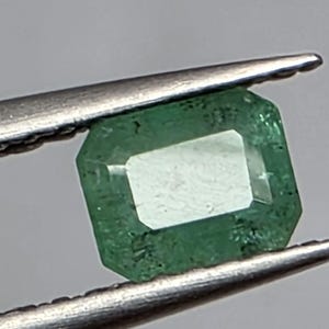 May include: A rectangular emerald-cut gemstone with a vibrant green hue and visible internal inclusions, held securely between the two metal tips of a pair of silver-toned tweezers. The stone features faceted edges that reflect light.