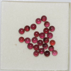 Può includere: Una collezione di 20 pietre preziose rotonde di colore rosso, probabilmente rubini, in una scatola bianca. Le pietre preziose sono disposte in un motivo a forma di cuore sciolto.
