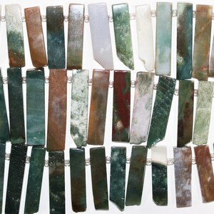 Peut inclure: Gros plan sur un fil de perles de pierres précieuses rectangulaires. Les perles sont un mélange de couleurs, dont le vert, le marron, le blanc et le rouge. Les perles sont enfilées avec un cordon clair et fin. Les pierres précieuses ont un aspect naturel et organique.