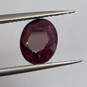 Peut inclure: Une pierre précieuse taillée en ovale, de couleur rouge-violet foncé, tenue par une pince. La gemme présente des facettes et des inclusions internes. L'arrière-plan est blanc uni. La pince est en métal argenté.