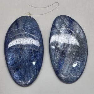 May include: Two oval-shaped, dark blue stones with a reflective, marbled appearance. The stones have a polished surface with streaks of lighter blue and white. A clear plastic loop is attached to one stone, suggesting they are intended for jewelry.