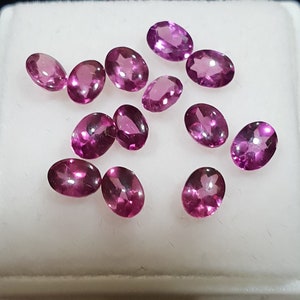 May include: A collection of oval-cut, vibrant pink gemstones. The gems are displayed on a white surface, showcasing their polished facets and rich color. The gemstones are arranged in a scattered pattern.