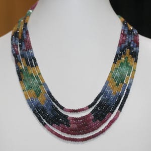 May include: A multi-strand gemstone necklace featuring a variety of colors including blue, green, yellow, and red. The necklace is displayed on a white mannequin and has a layered design, with each strand showcasing a different hue.
